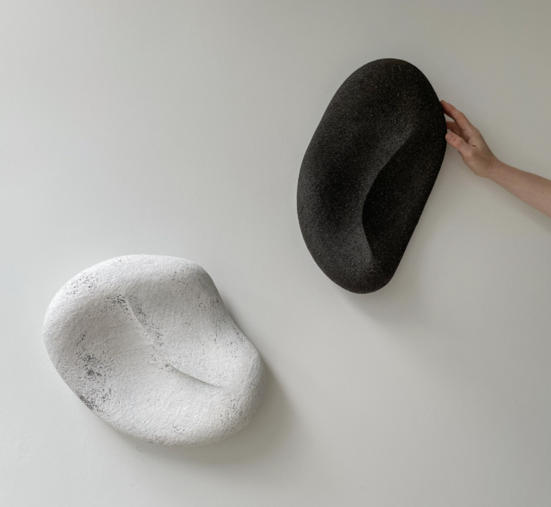 'Taurus No. 26' by Yasha Butler features two wall-mounted ceramic sculptures: one in matte black and one in chalk white. Both pieces have smooth, organic forms reminiscent of river stones, with gentle curves and indentations. A hand reaches into frame to touch the black sculpture, emphasizing its scale and tactile quality. The pieces are displayed against a white wall, creating a striking contrast with the dark sculpture.